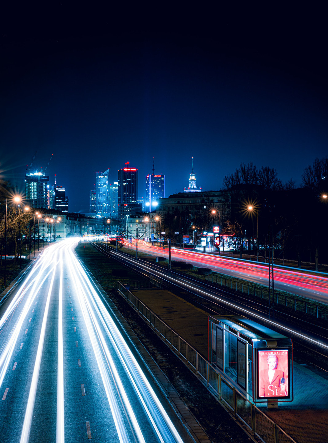 Langzeitbelichtung einer viel befahrenen Straße bei nacht, im Hintergrund die Skyline einer modernen Stadt